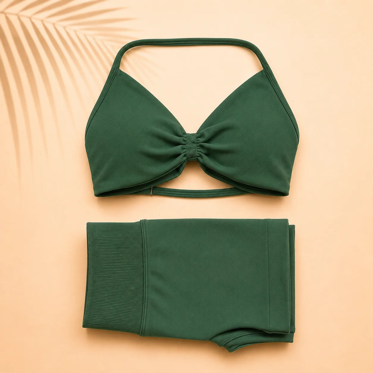 A two-piece dark green swimwear set consisting of a halter-neck bikini top with a front twist detail and matching high-waisted bottoms, laid flat on a peach background with a palm leaf shadow.