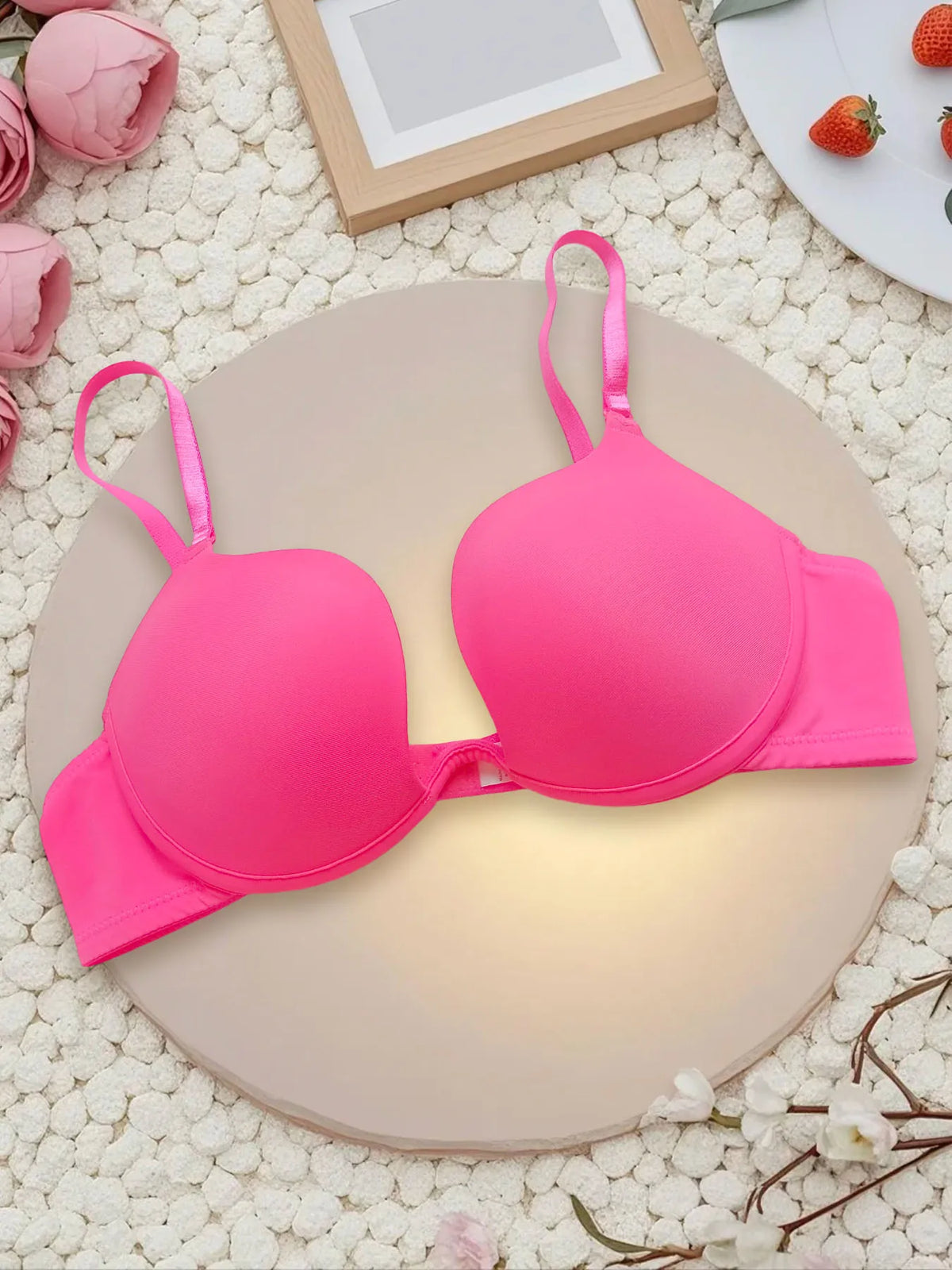 A bright pink wireless underwire bra displayed on a round beige platform, surrounded by decorative props including pink flowers, white pebbles, a wooden frame, and strawberries.