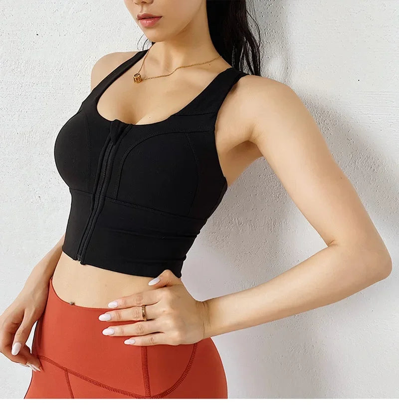 A woman wearing a black athletic sports bra with a front zipper and structured cups, paired with rust-orange leggings, standing against a light textured wall.