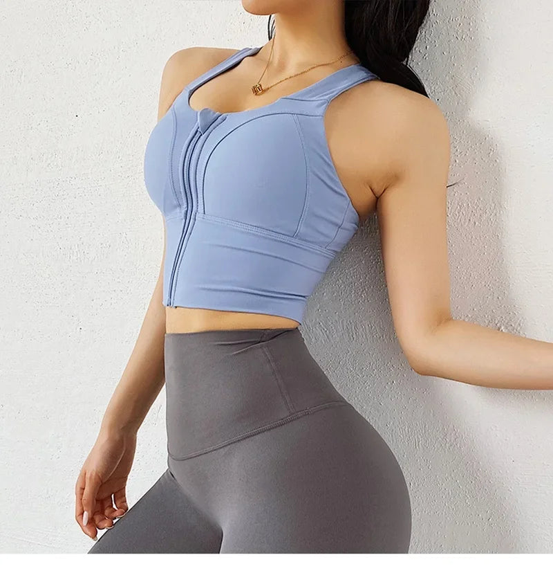 A woman wearing a light blue sports bra with a front zipper and high-cut gray leggings, leaning against a textured white wall.