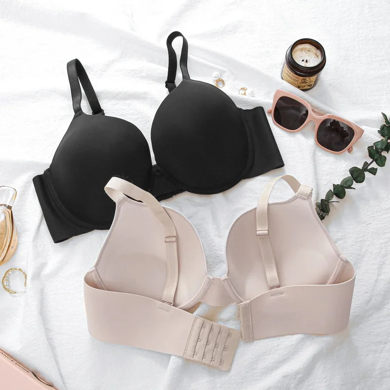Two wireless underwire bras arranged on a white fabric background, one in black and one in light blush beige. Pink sunglasses, a candle, rings, and a small green sprig are placed around them as styling props.