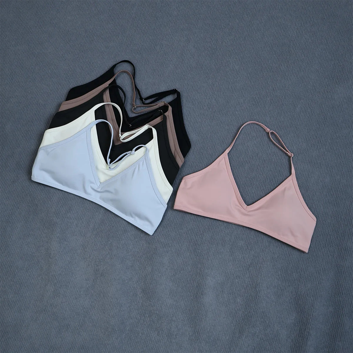 Three minimalist sports bra or bikini top styles arranged on a textured gray-blue fabric background. The set includes a light blue top, a white top with black side panels and brown trim, and a separate light pink top.
