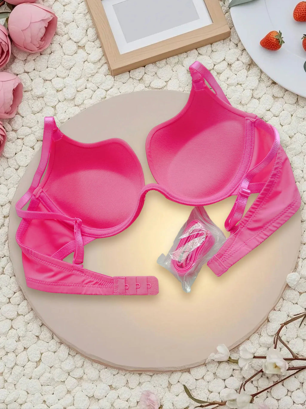 A bright pink bralette set with matching underwear and a small packaged accessory, arranged on a round beige board over a white textured surface with decorative flowers and strawberries.