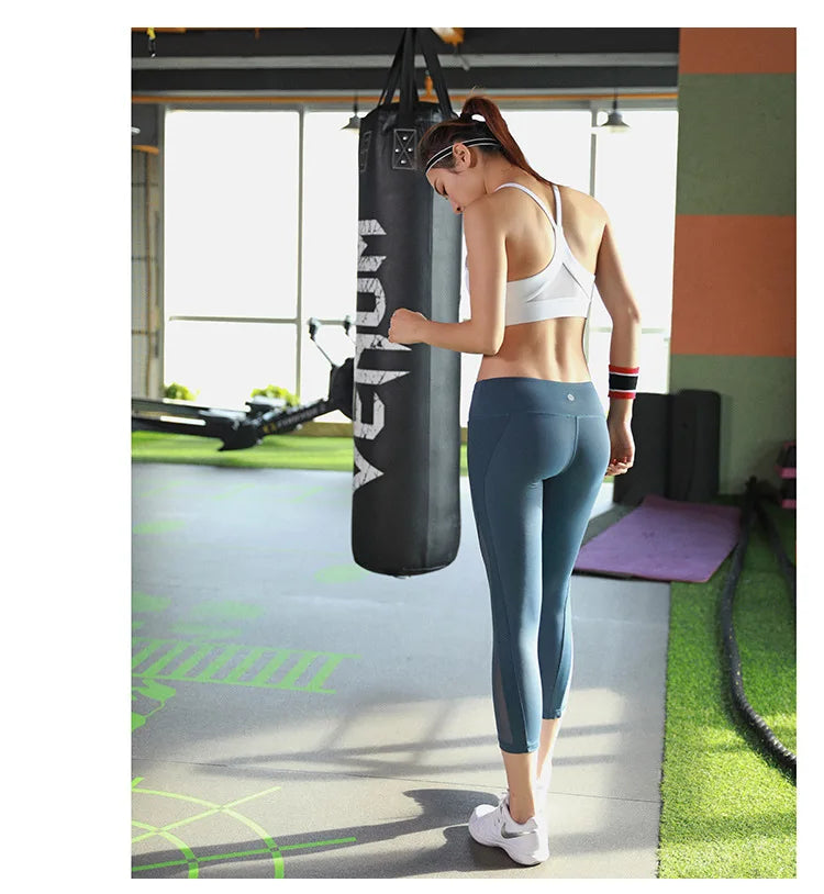 A woman in a gym wearing a white sports bra and blue leggings, standing next to a black punching bag.