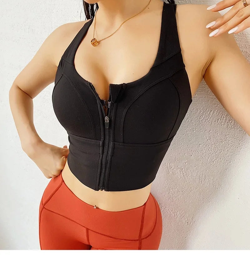 A woman wearing a black athletic bustier-style sports bra with a front zipper and orange-red fitted leggings, photographed against a light textured wall.