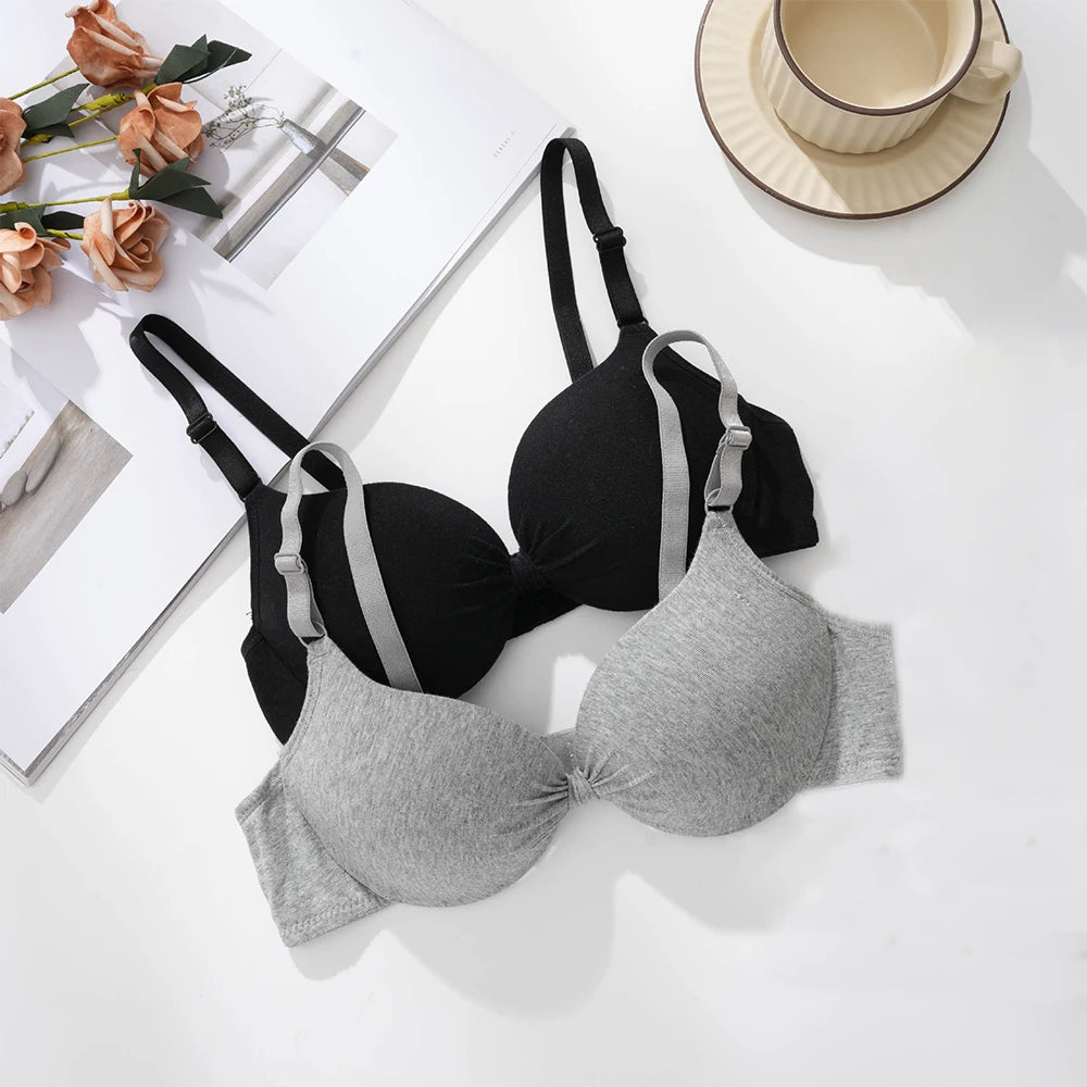 Two wireless underwire bras laid flat on a white surface: one black with light gray side panels and straps, and one heather light gray. Decorative flowers, a magazine page, and a cup on a saucer are placed around them.