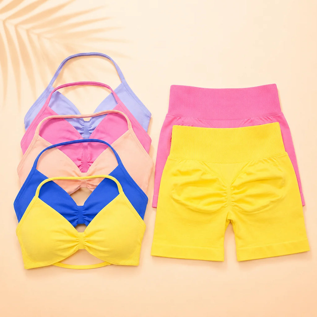 Four women's activewear pieces arranged flat on a warm beige background: two bra tops in blue, pink, peach, and lavender, and two shorts in pink and bright yellow. No person, animal, or real environment is present, so this is Product/Packaging.