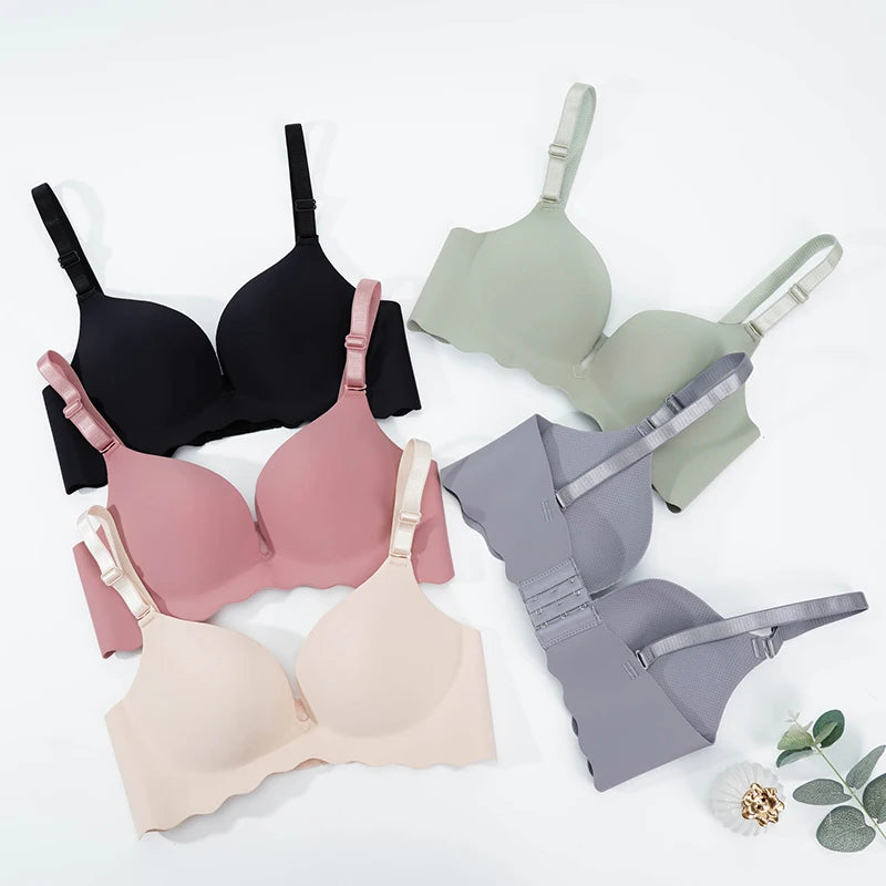 Four wireless underwire bras in different colors are arranged in a flat-lay on a white background: black, sage green, dusty pink, and light beige, with one gray bra shown from the back. A small decorative flower and eucalyptus leaves sit in the lower right corner.