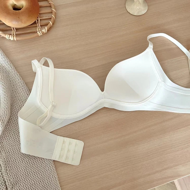 A white wireless underwire bra laid flat on a light wood tabletop, with a matching white hook-and-loop back strap piece placed beside it. A woven tray with an apple and a small candle are partially visible in the background as styling props.