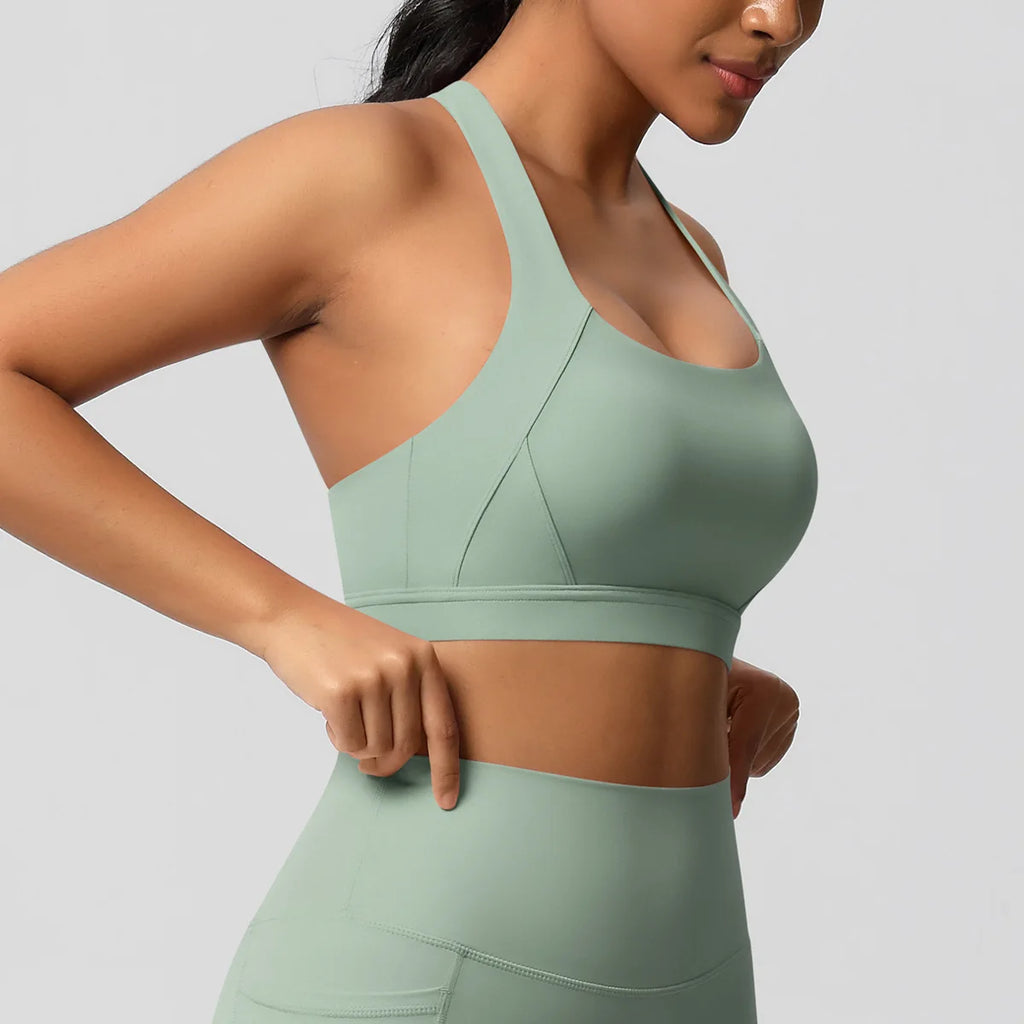 A woman wearing a sage green sports bra and matching high-waisted leggings against a solid light gray background.