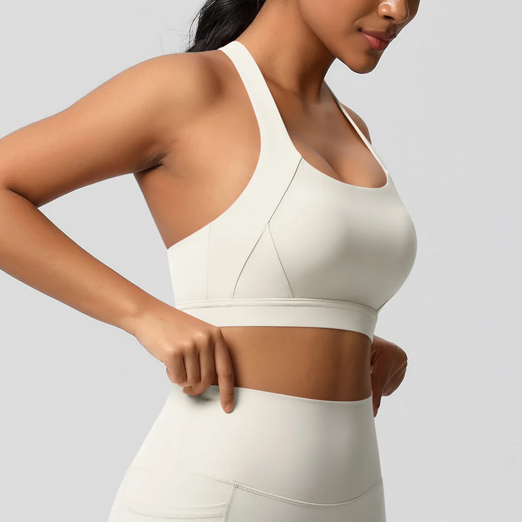 A close-up of a woman wearing a light cream/off-white sports bra and matching high-waisted leggings against a solid light gray studio background.