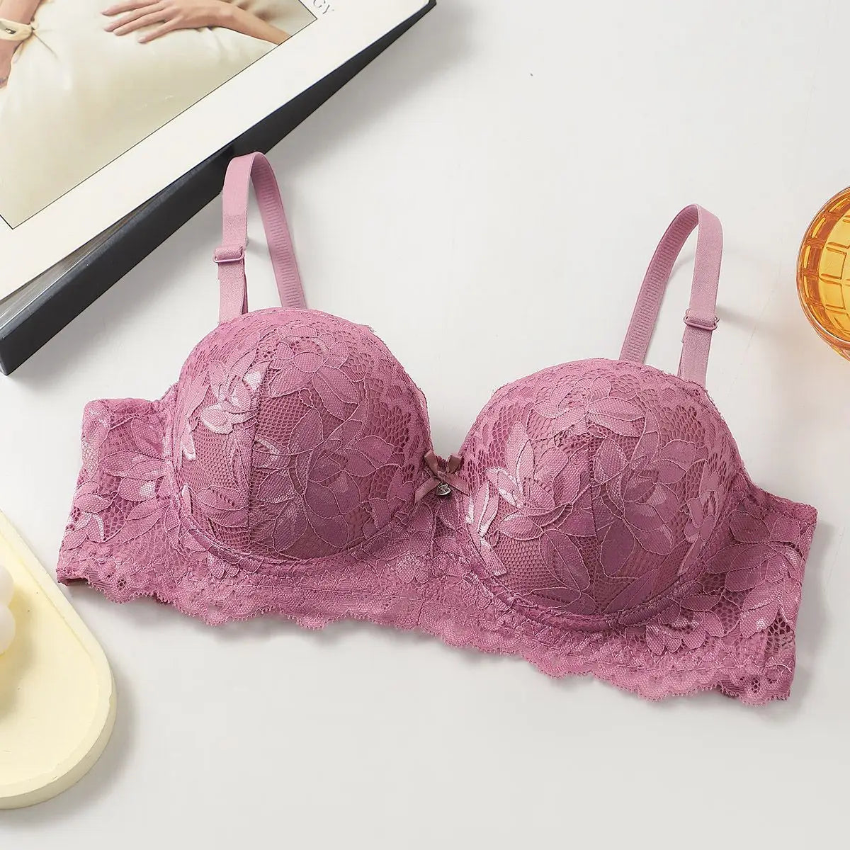 A dusty pink floral lace underwire bra laid flat on a white surface, with a magazine page and a small yellow object partially visible as styled props.